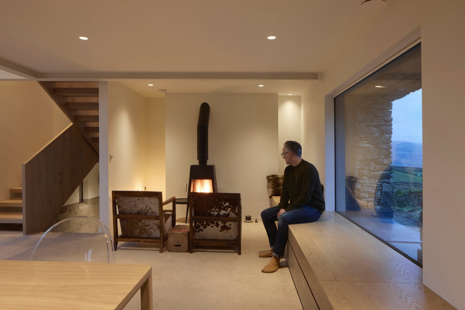 Cosy barn lounge at dusk with log burner, modern oak staircase, and large glazed window with fitted window seat
