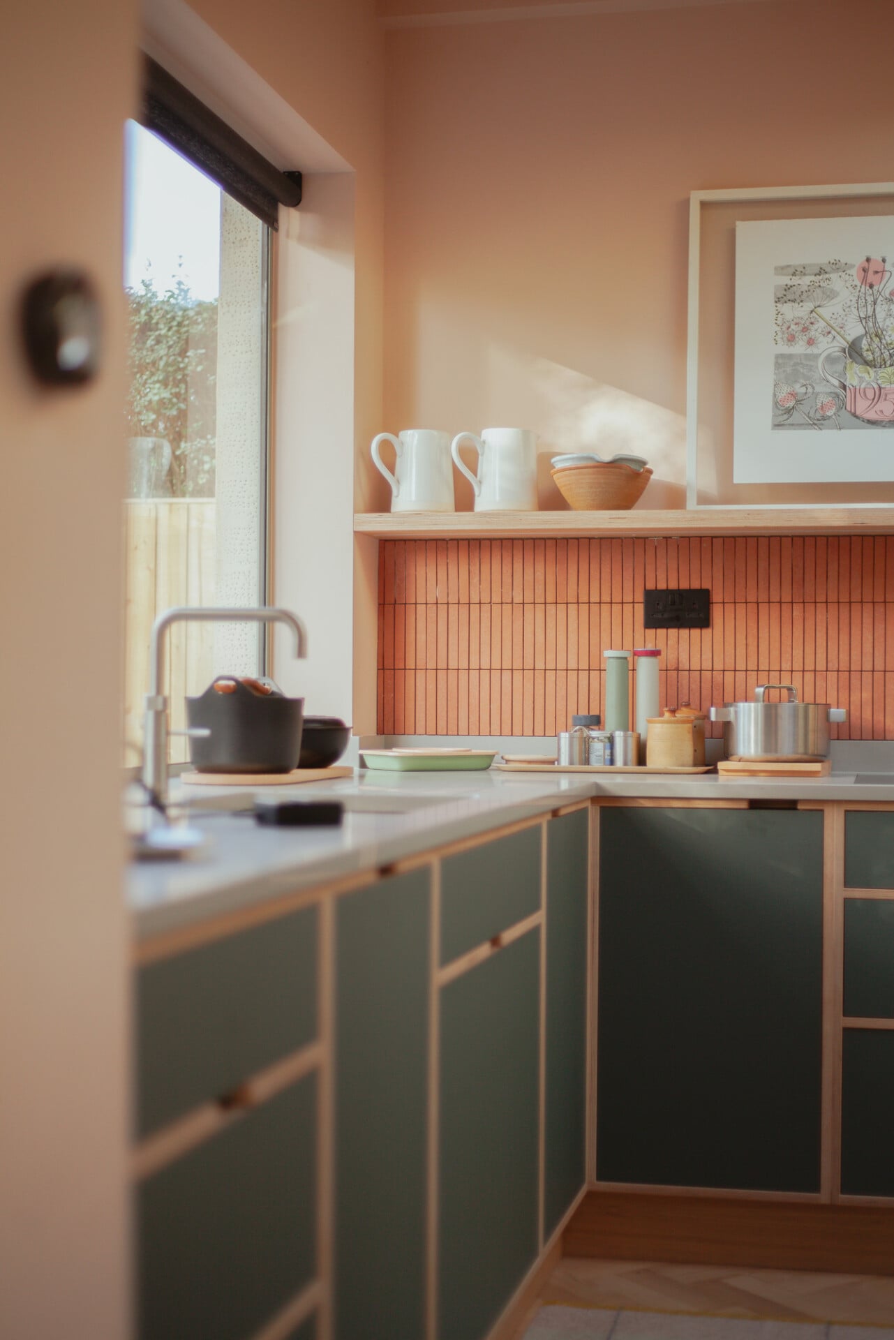 Soft pink kitchen with green cabinets, pink tile splashback, and open wood shelving in London