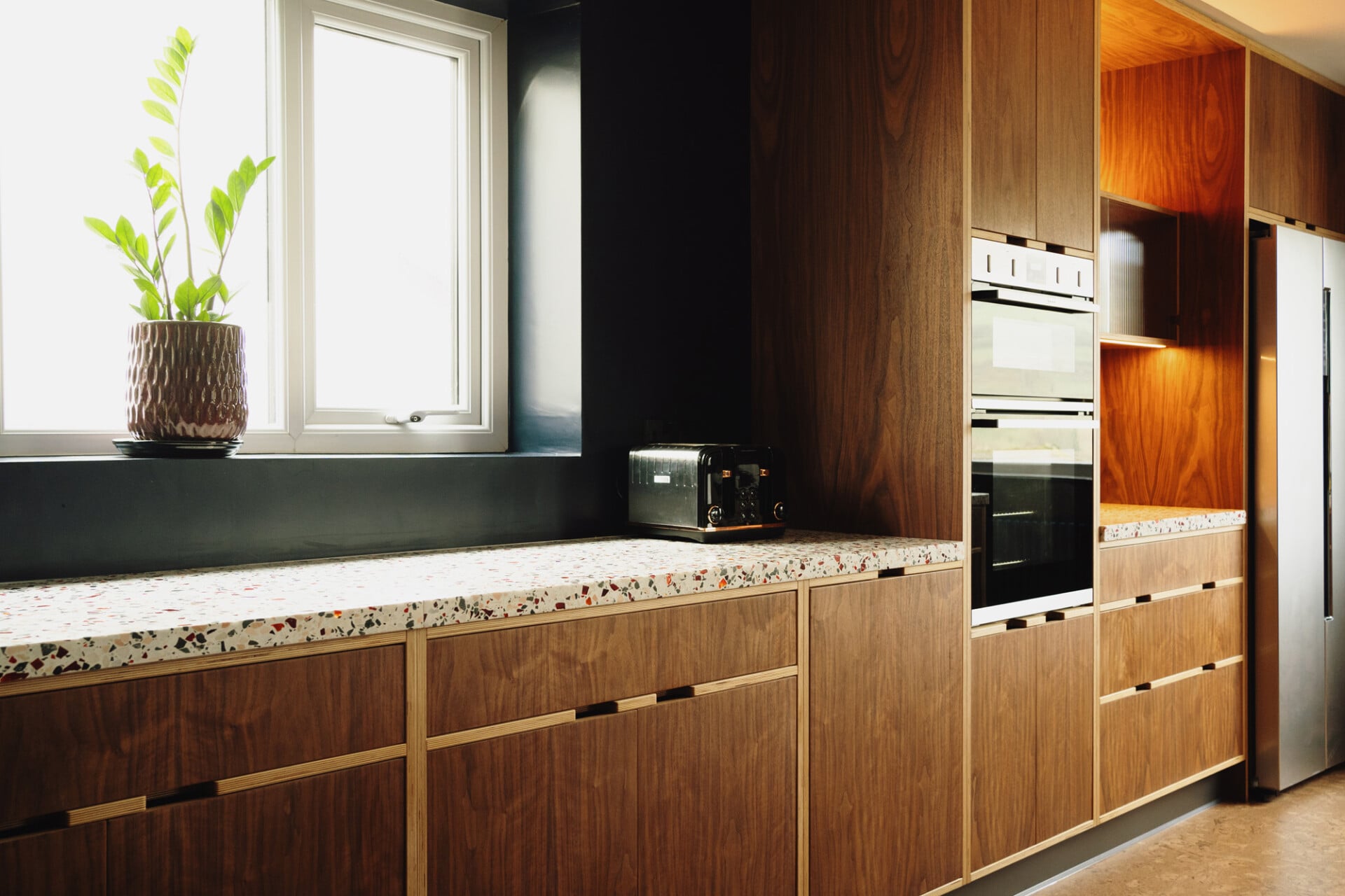 Modern walnut plywood kitchen with terrazzo worktop, tall units, and black walls