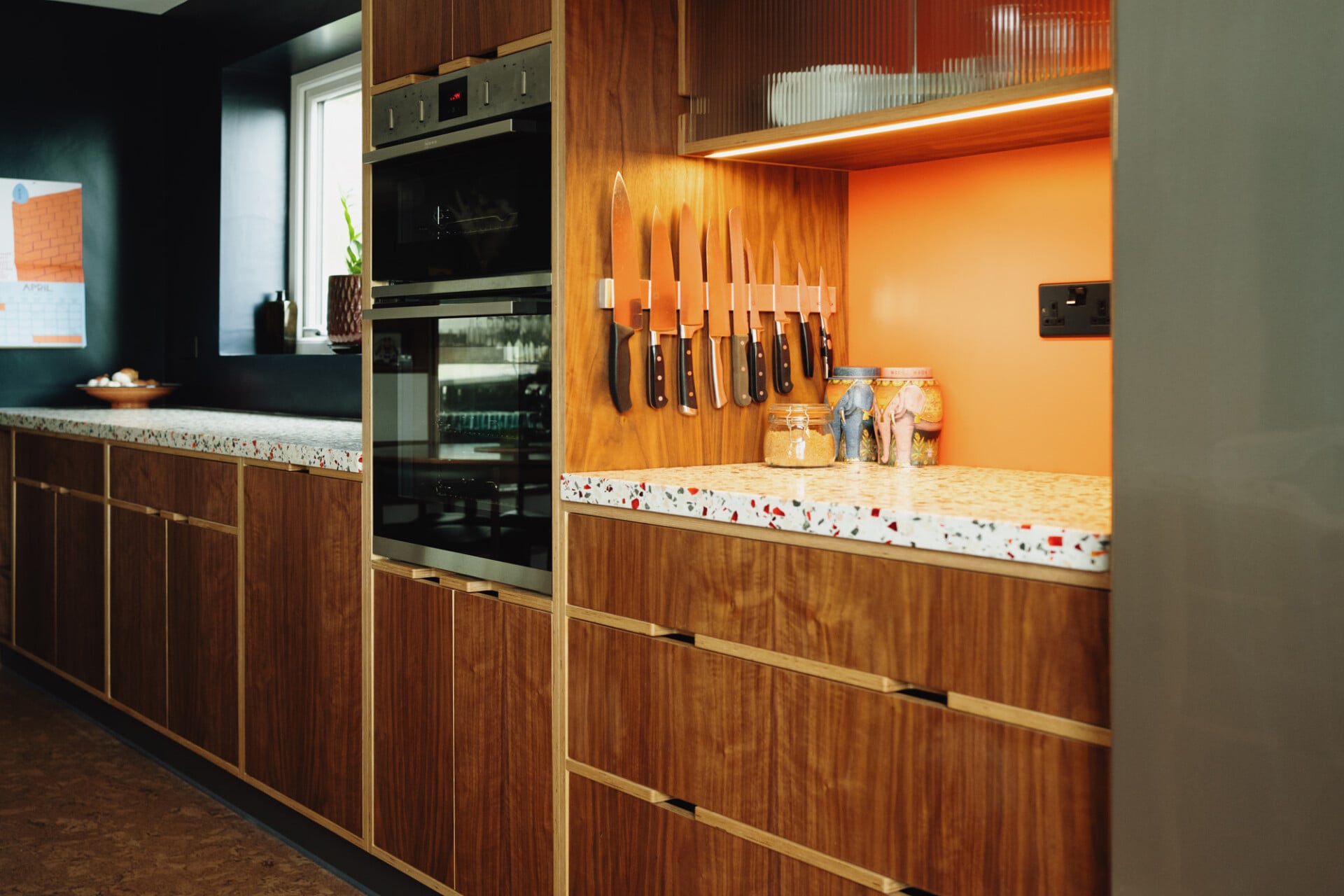 Walnut plywood kitchen with orange splashback, terrazzo counters, and open shelving
