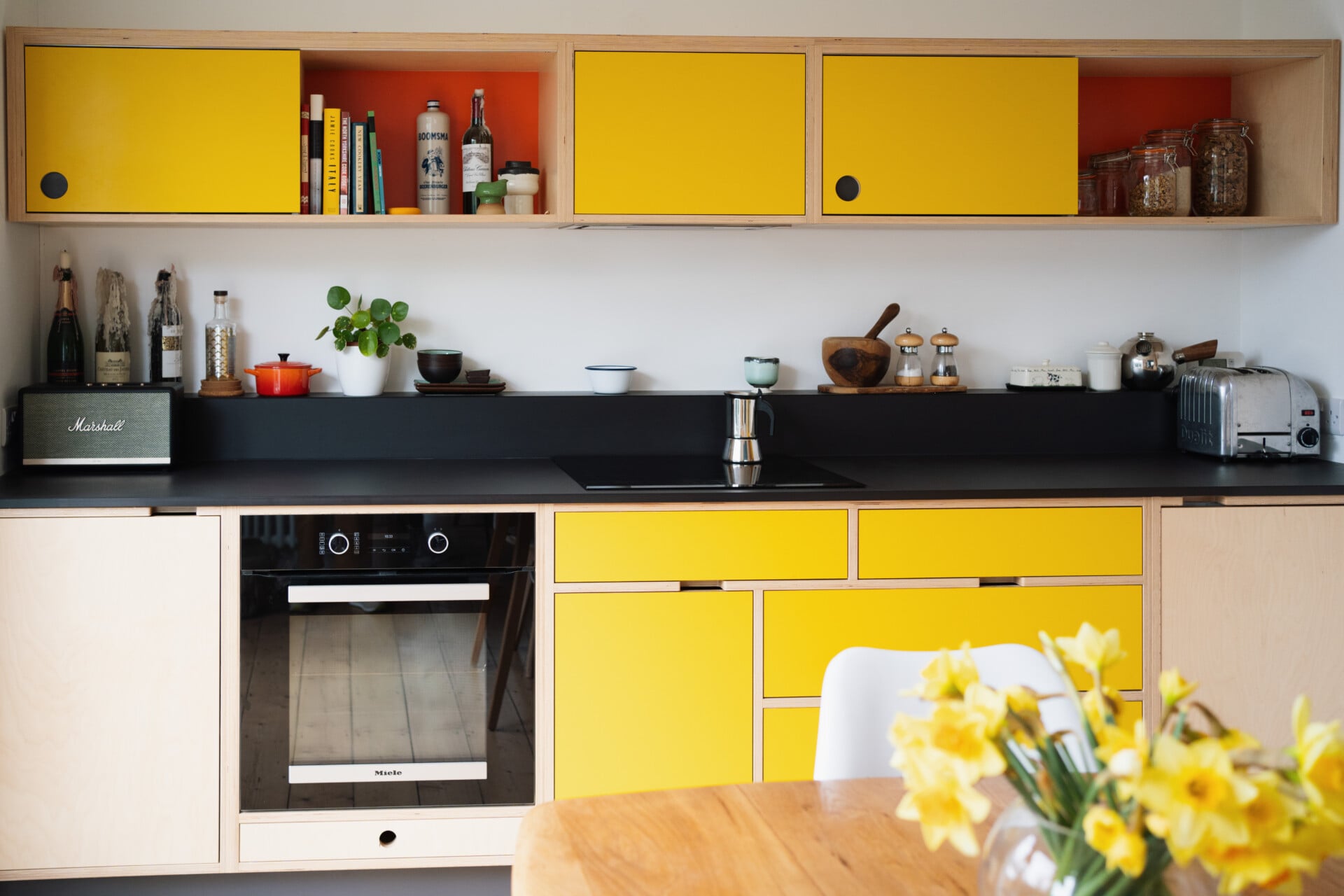 Yellow plywood kitchen with black splashback and worktops, open shelving, and integrated oven